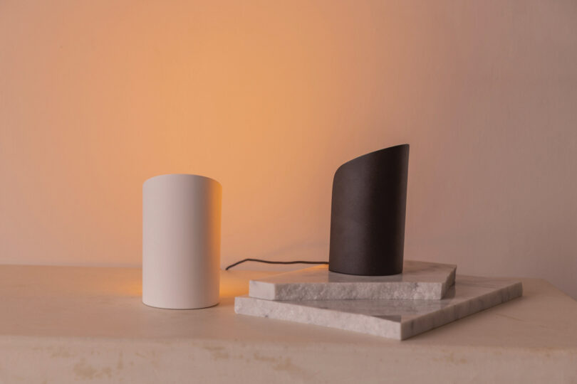 A white cylindrical object and a dark, slanted cylindrical object are placed on stacked marble slabs against a softly lit beige background.