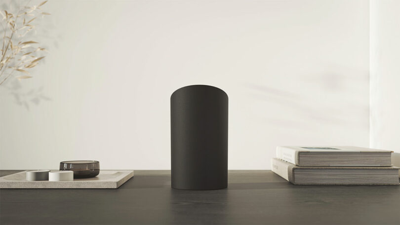 A black cylindrical object with a deep hue is placed on a dark tabletop between a stack of books and decor items, against a light-colored wall.