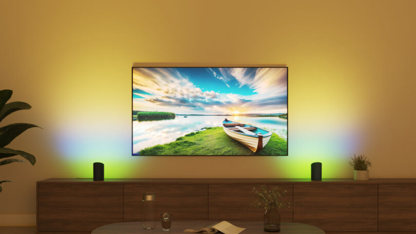 A wall-mounted TV displays a scenic lake image with ambient backlighting. Below, a wooden console holds speakers, a glass, a vase with flowers, and decorative items.