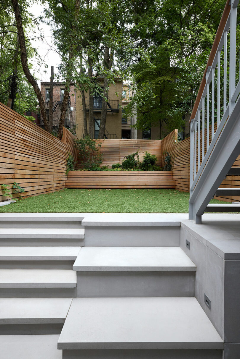 Modern backyard with white concrete steps leading to a grassy area, wooden privacy fencing, planters with shrubs, and surrounding trees.