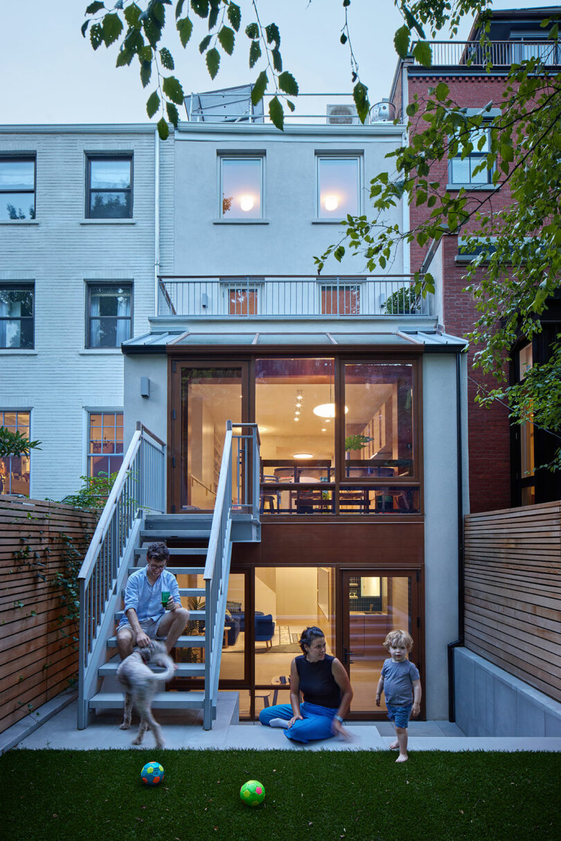 A family with a dog and child spends time in a modern backyard with artificial turf, colorful balls, and a multi-story urban townhouse in the background.