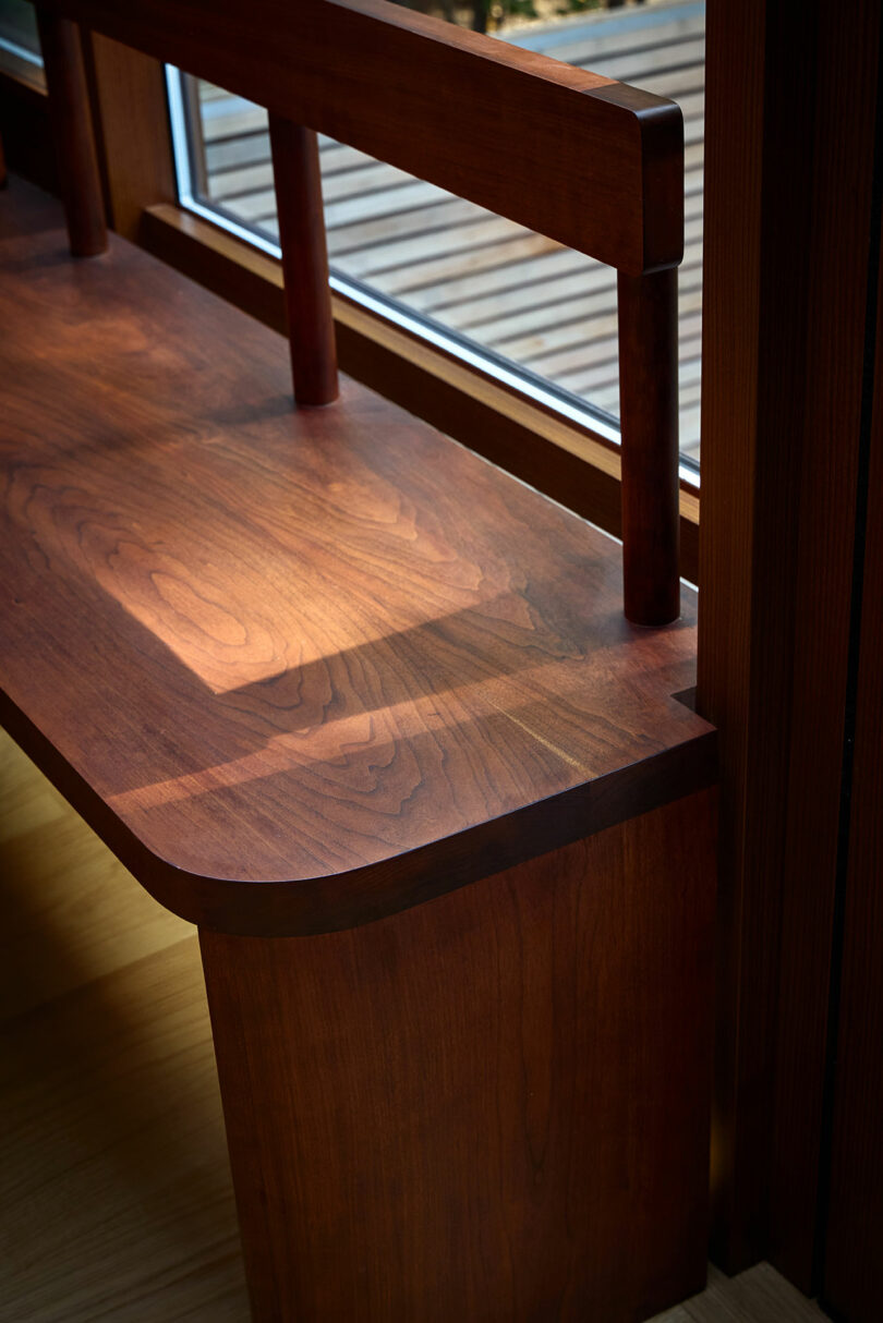 A wooden bench with a backrest is positioned next to a window, casting shadows on its surface from natural light.