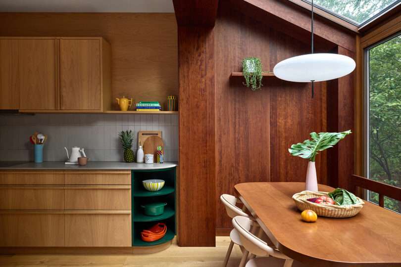 Modern kitchen with wooden cabinets, open shelving, a round dining table with chairs, a skylight, and a basket of fruits and vegetables on the table.