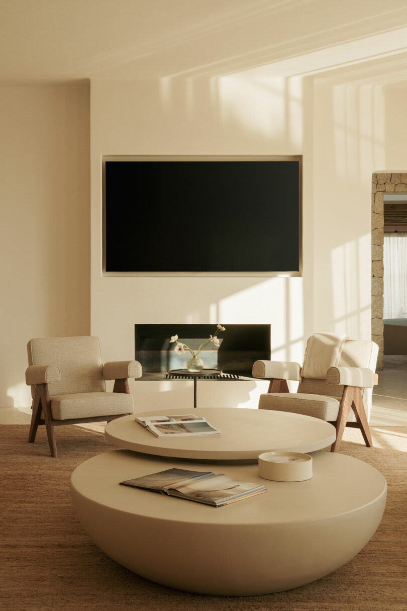Minimalist living room with two beige armchairs, round beige coffee tables, a blank wall-mounted TV, fireplace, and magazines on the tables. Neutral tones and natural sunlight.