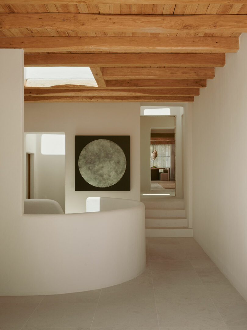 Minimalist interior with smooth white walls, light tile floor, exposed wooden ceiling, and a large circular artwork on the wall near white stairs.