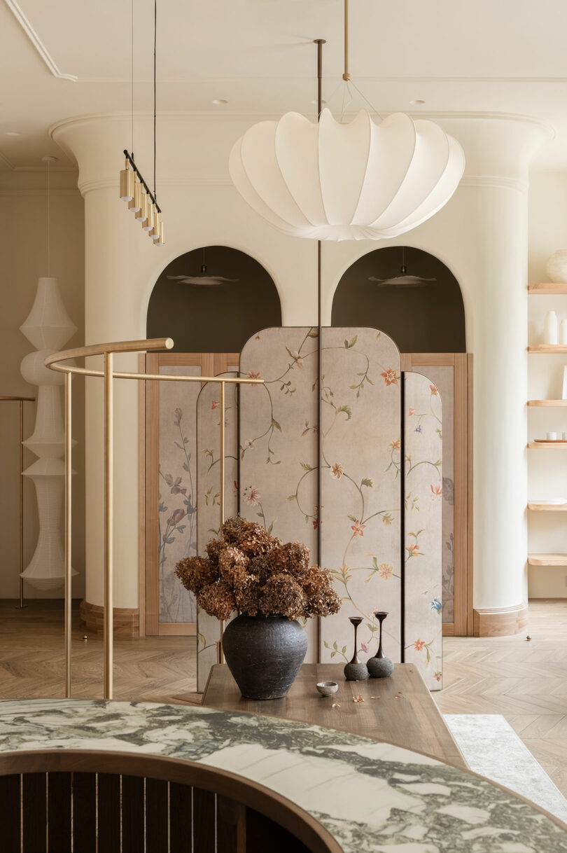 A modern interior featuring a marble-topped counter, a wooden table with dried flowers, a floral room divider, and minimalist lighting fixtures.