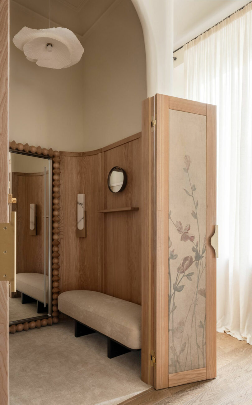 A small dressing area with light wood paneling, a beige upholstered bench, a folding screen with floral design, a round mirror, and soft natural light through sheer curtains.