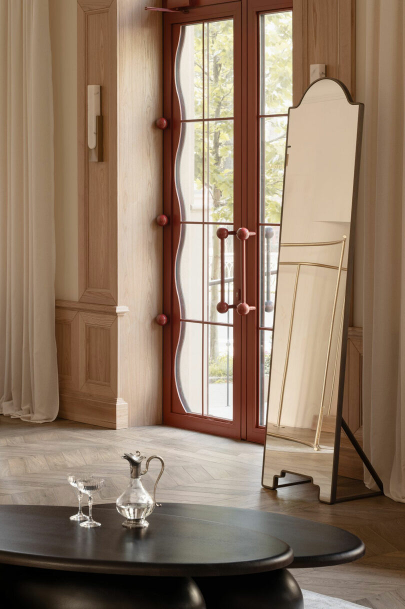 A full-length mirror stands near red-framed glass doors in a room with light wood walls, sheer curtains, and a dark coffee table set with a decanter and two glasses.