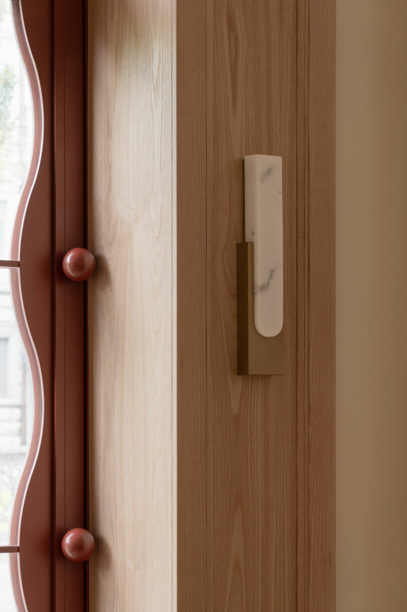 A modern wall sconce with a marble accent is mounted on a light wood wall next to a red-framed window with round decorative knobs.