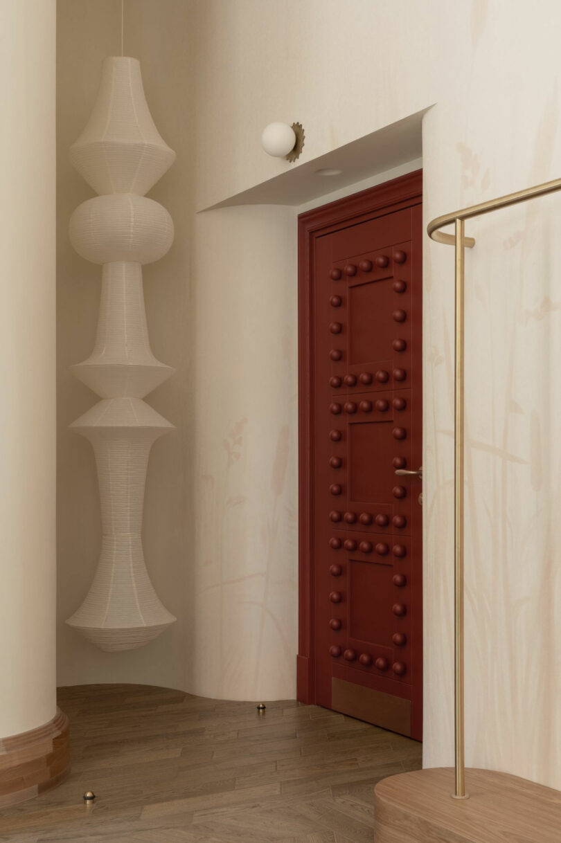 A red door with raised circular knobs is set in a neutral hallway with light wooden floors, a tall sculptural white lamp, and a gold clothing rack.