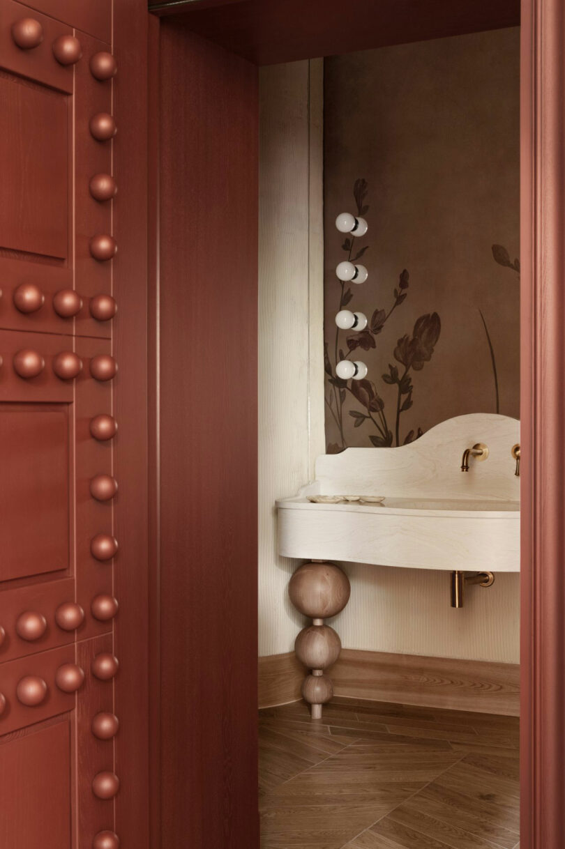 A view through a red door reveals a bathroom with a white ornate sink, brass faucets, floral wall art, and wood-patterned flooring.