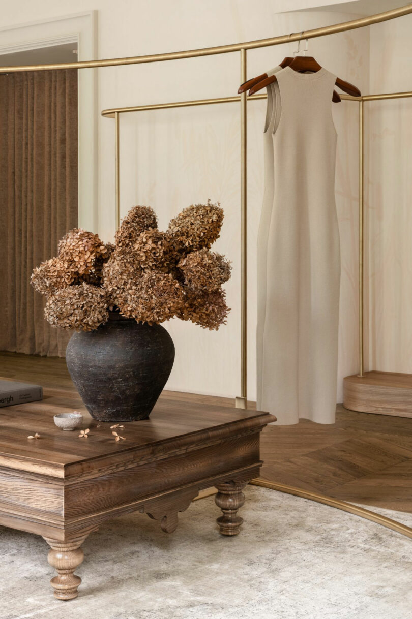 A sleeveless beige dress hangs on a brass clothing rack behind a rustic wooden table with a black vase of dried hydrangeas.