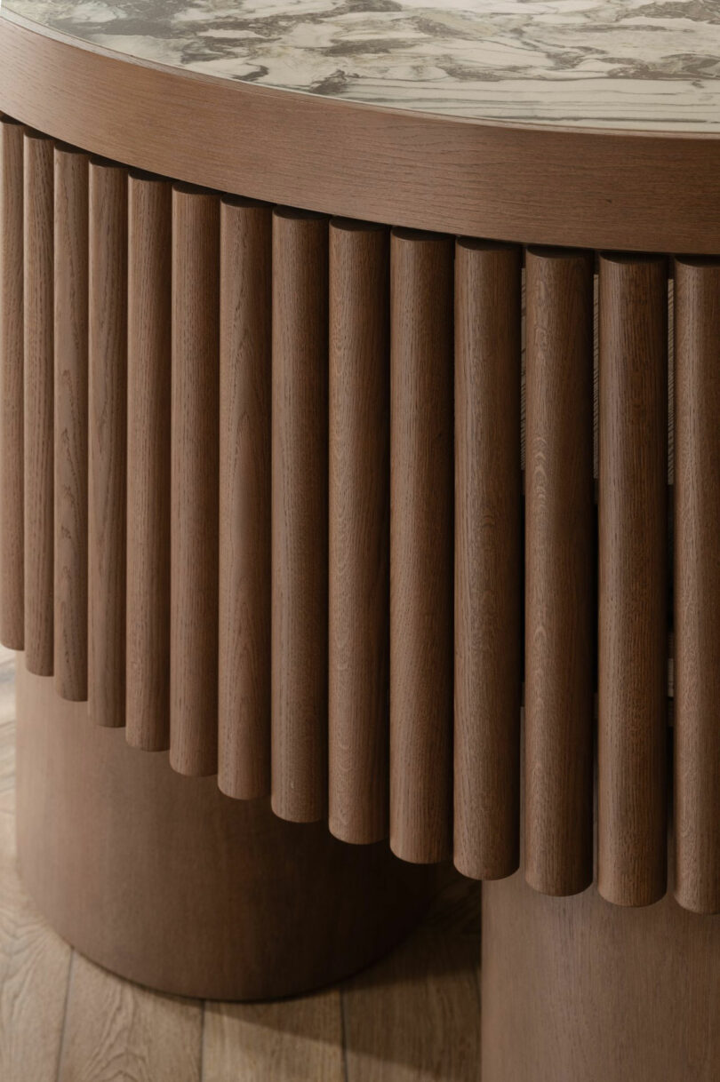 Close-up of a wooden table base with vertical cylindrical slats and a round marble tabletop on a wooden floor.