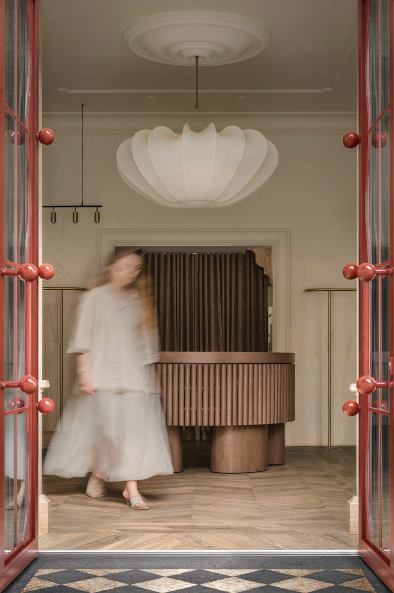 A blurred person in light clothing walks inside a room with wood furniture, a large pendant light, and red-framed double doors.
