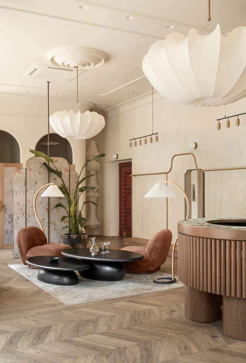 Modern lounge with curved brown chairs, round black tables, potted plant, large white pendant lights, and a wooden reception desk on herringbone wood flooring.