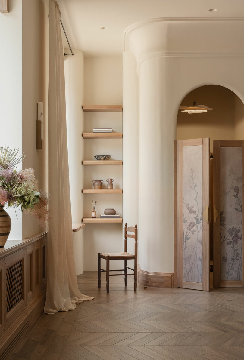 Minimalist interior with wooden chair, open shelves displaying ceramics, arched doorway, folding screen with floral design, soft beige walls, and a vase of flowers on the left.