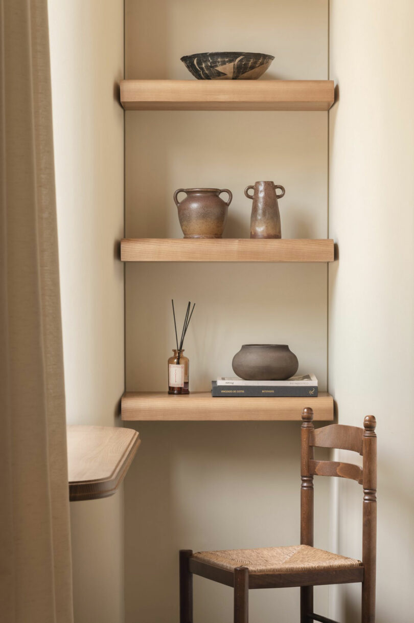 Three wooden shelves display ceramic vessels, a bowl, a reed diffuser, and books in a minimalist, neutral-toned room with a wooden chair and a table partially visible.