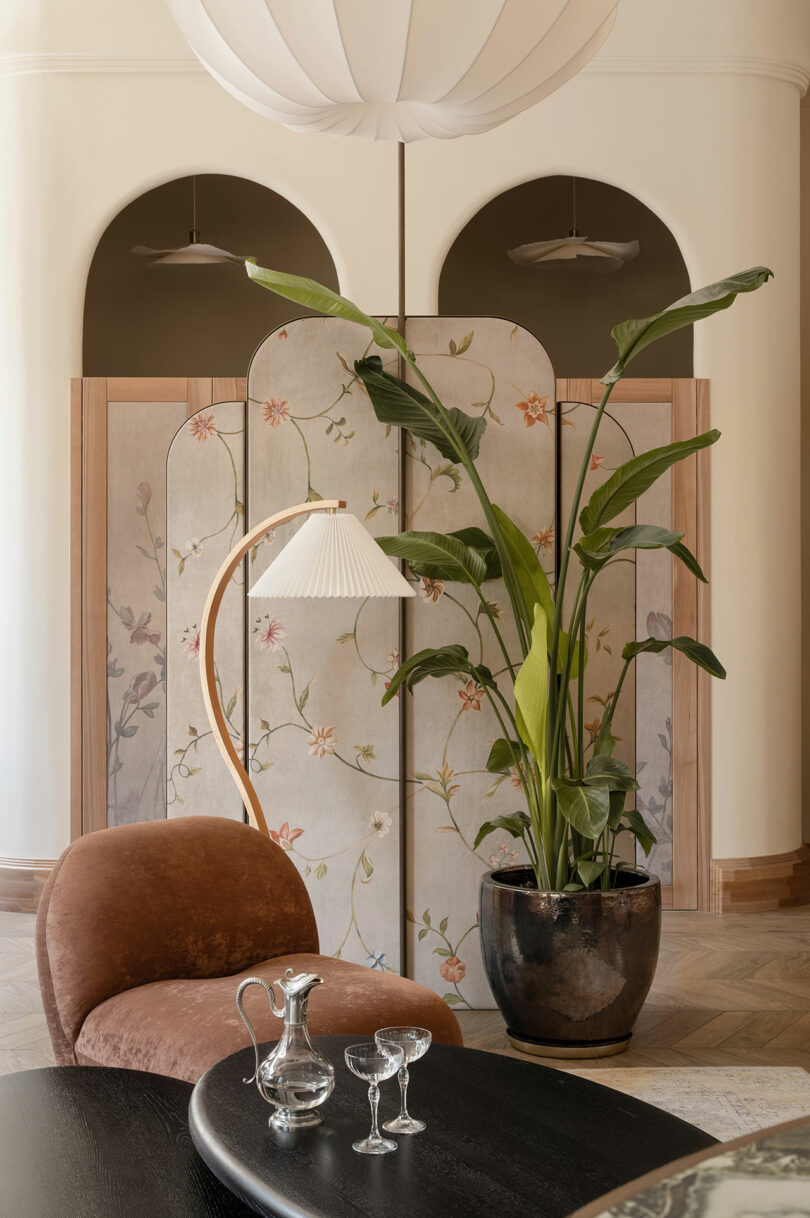 A modern living room with a curved brown lounge chair, a floor lamp, a black coffee table with glassware, a large potted plant, and a floral folding screen in the background.