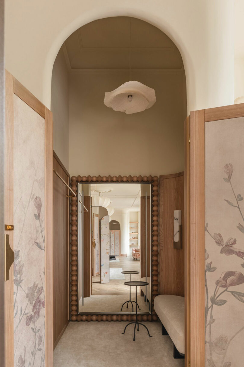 A hallway with floral-patterned panels, a wooden-framed mirror, a cushioned bench, a small round table, and a sculptural ceiling light.