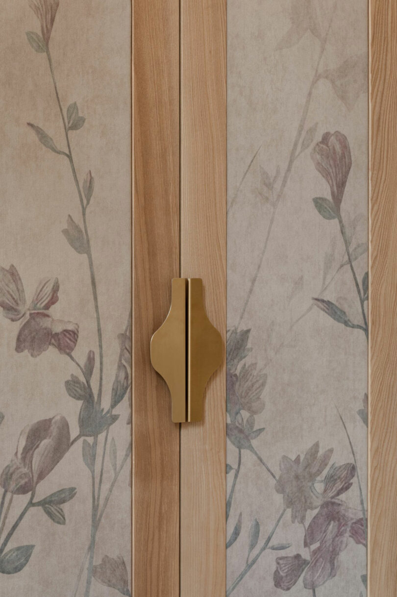 Close-up of wooden cabinet doors with a central brass handle and floral-patterned wallpaper featuring muted pink and green tones.