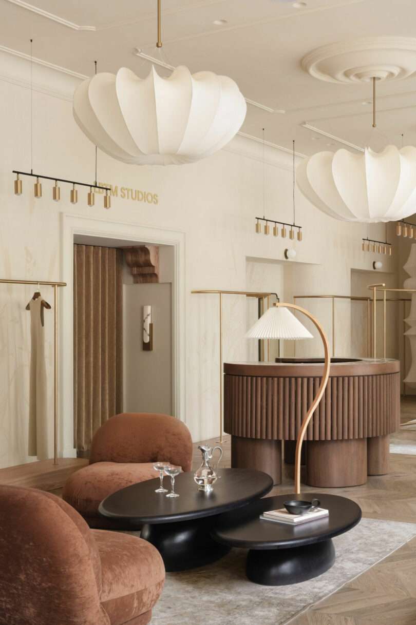 Modern lounge with curved wooden reception desk, sculptural pendant lights, two round brown chairs, black nesting tables, and minimal decor in neutral tones.
