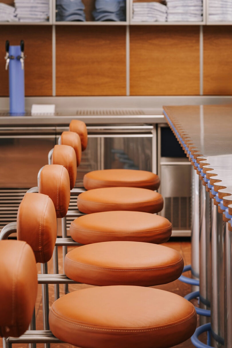 Row of round, orange cushioned bar stools lined up along a metallic counter in a modern restaurant, with shelves and folded towels in the background—giving the space a sleek, Pluto-inspired vibe.