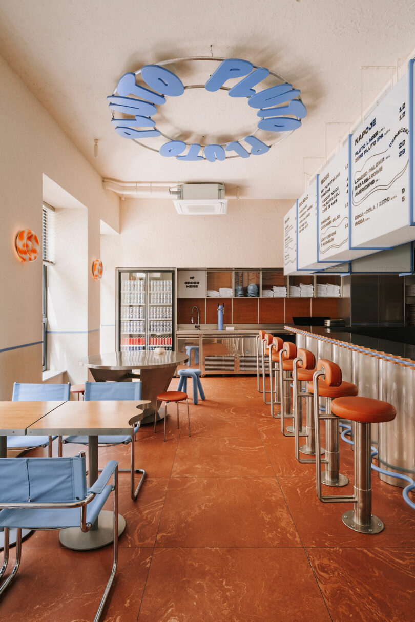 Modern café interior with blue and orange decor, metal bar stools, tables with chairs, a menu board overhead, and a fridge stocked with cans in the back—Pluto’s inspired charm is felt throughout.