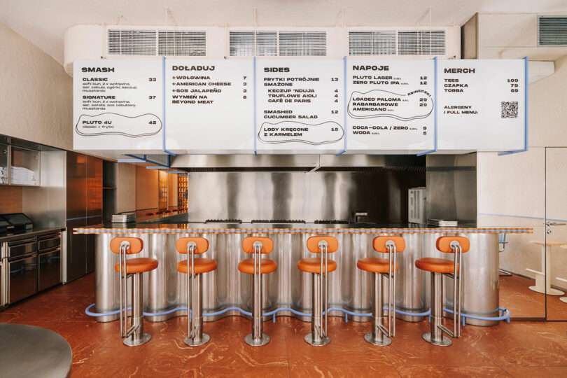 Modern diner interior with metallic counter, six orange bar stools, stainless steel appliances, and large overhead menu signs displaying food, drink, Pluto-inspired treats, and merchandise options.
