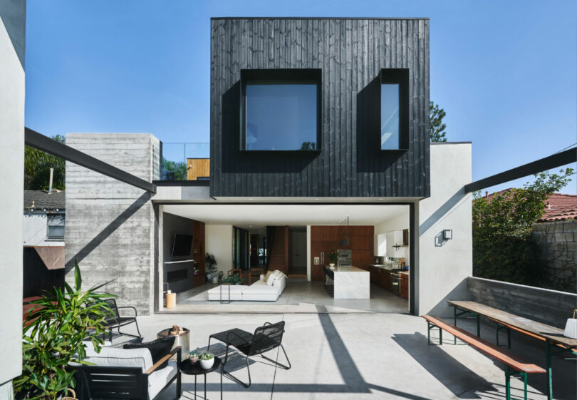 Modern two-story house with an open living area, concrete and wood finishes, large windows, and outdoor seating under a clear blue sky.