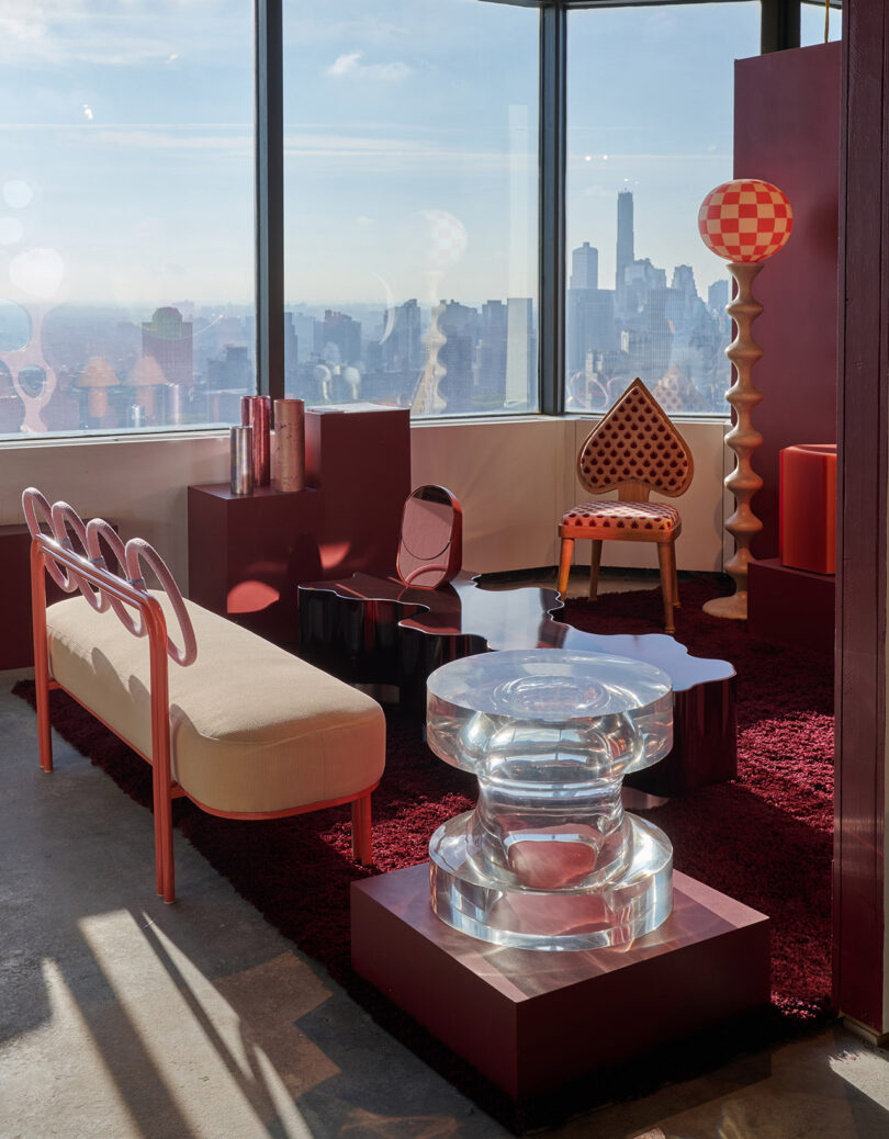 A modern room with eclectic red and beige furniture, a sculptural clear table, and large windows overlooking a city skyline.