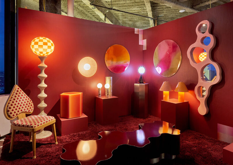 A room with red walls and carpet features eclectic furniture, round mirrors, geometric lamps, and a sculptural shelf, all in warm lighting and bold, colorful designs.