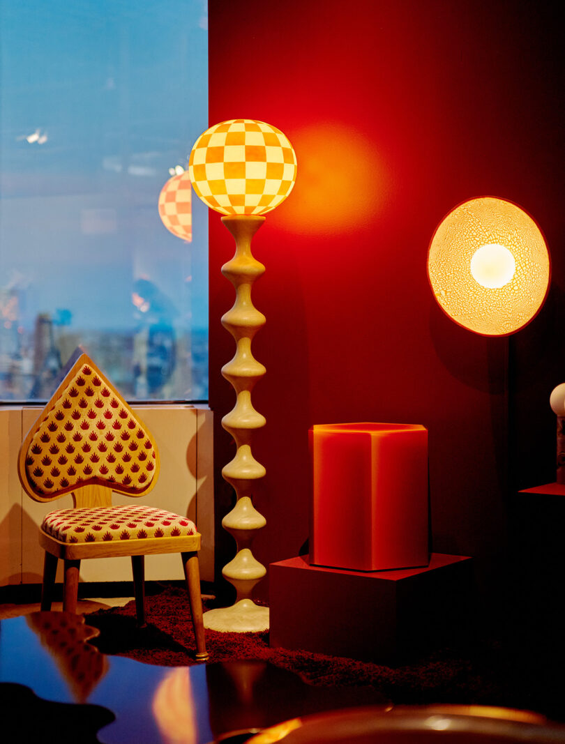 A room with a heart-patterned chair, a tall checkered floor lamp, a round wall light, and a large red cube-shaped seat against a dark red wall.