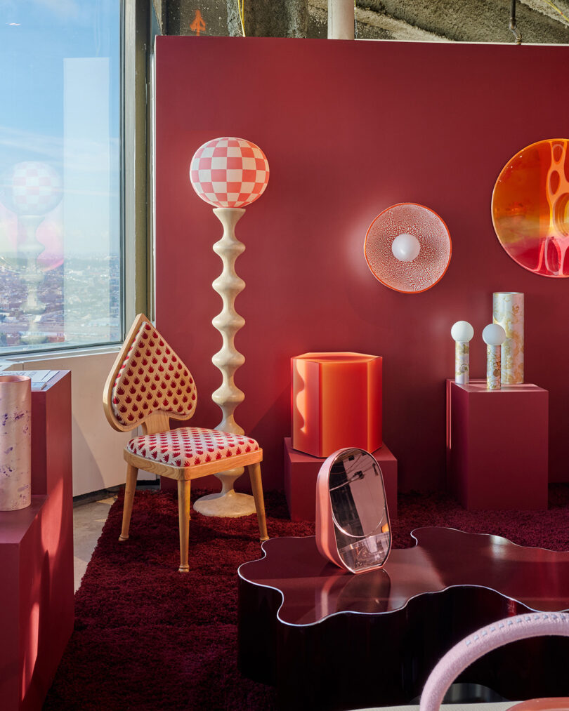 A modern interior with deep red walls and carpet, geometric furniture, a patterned lamp, circular mirror, and a heart-shaped chair by a large window.