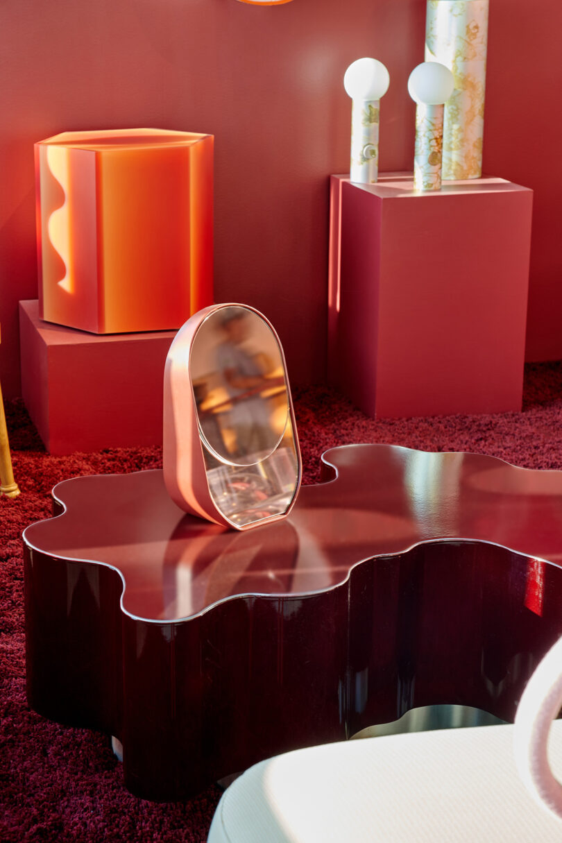 A sculptural mirror with a metallic pink frame sits on a wavy dark wood table, surrounded by geometric objects and red-toned decor.