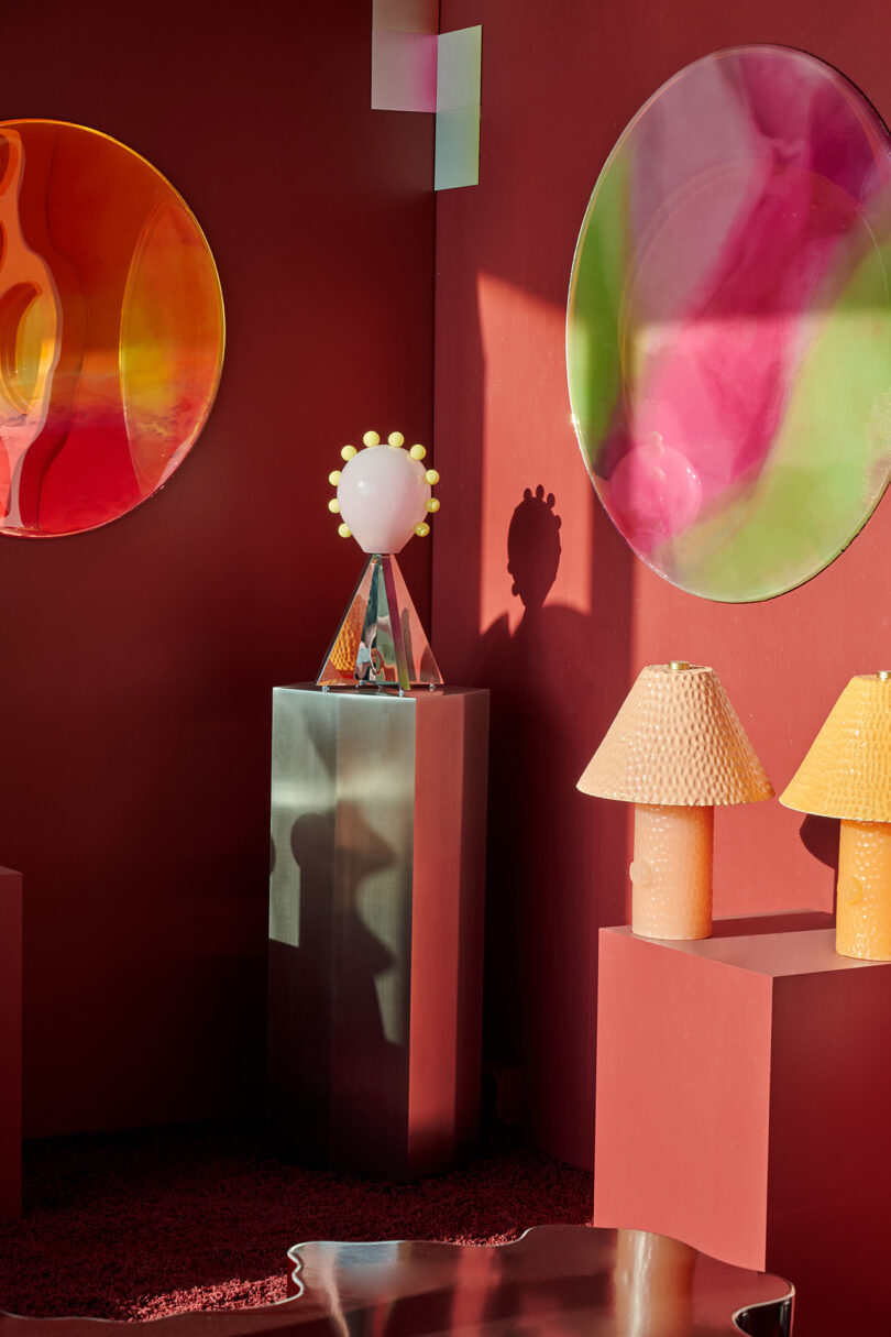 A modern interior with red walls features abstract circular wall art, a sculptural lamp on a pedestal, and two textured table lamps on a red surface.