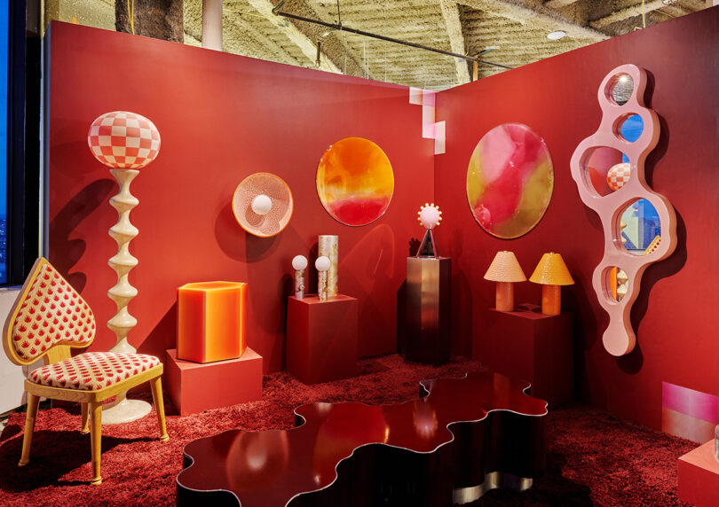 A room with deep red walls and carpet features bold, colorful modern furniture, sculptural lamps, patterned chairs, circular mirrors, and contemporary decorative objects.