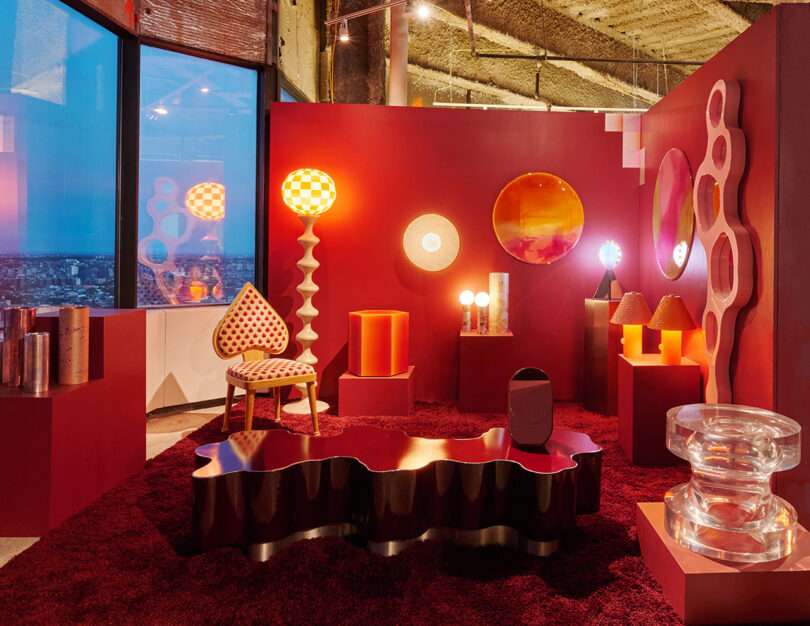 A modern room features bold red walls and decor, mirrored accents, geometric lamps, patterned furniture, and sculptural objects, with large windows overlooking a cityscape.