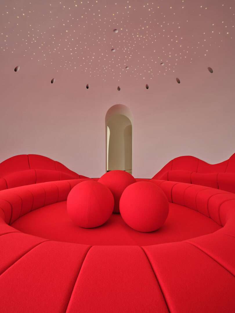 A modern room with curved red seating, large red spheres in the center, an arched doorway, and a ceiling with small recessed lights resembling stars.