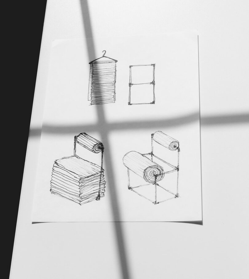 A sheet of paper with four black ink sketches showing chairs and furniture designs made from rolled and stacked towels on a white surface with a shadow crossing over it.