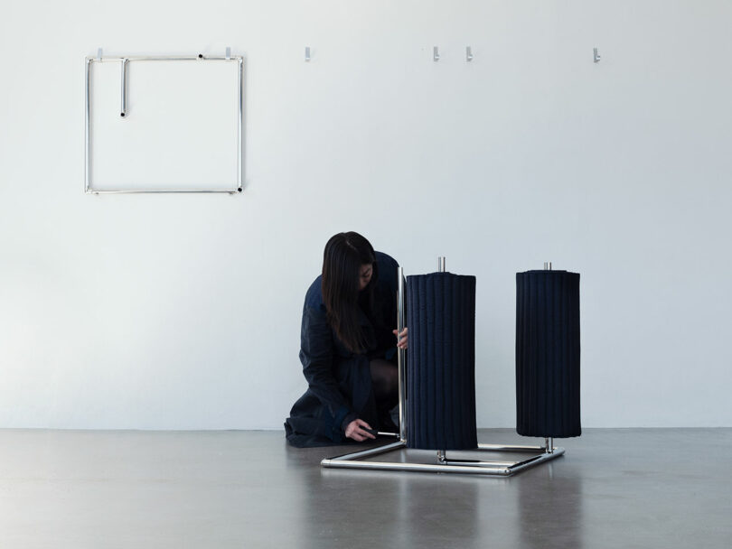 A person kneels on the floor assembling a metal and fabric structure in a minimalist room with a white wall and metal hooks.