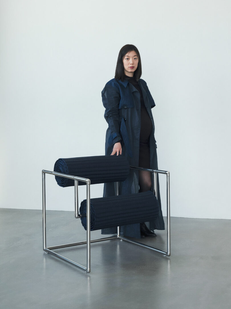 A woman in a long dark coat stands indoors next to a modern chair with a metal frame and rolled cushions, against a plain light-colored wall.