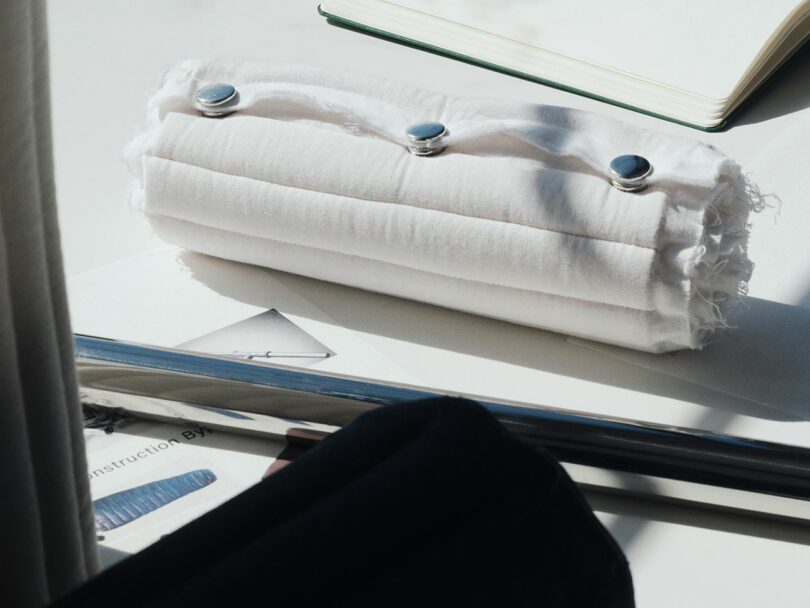 A cylindrical white fabric roll with three silver buttons sits on a desk next to an open notebook and some papers.