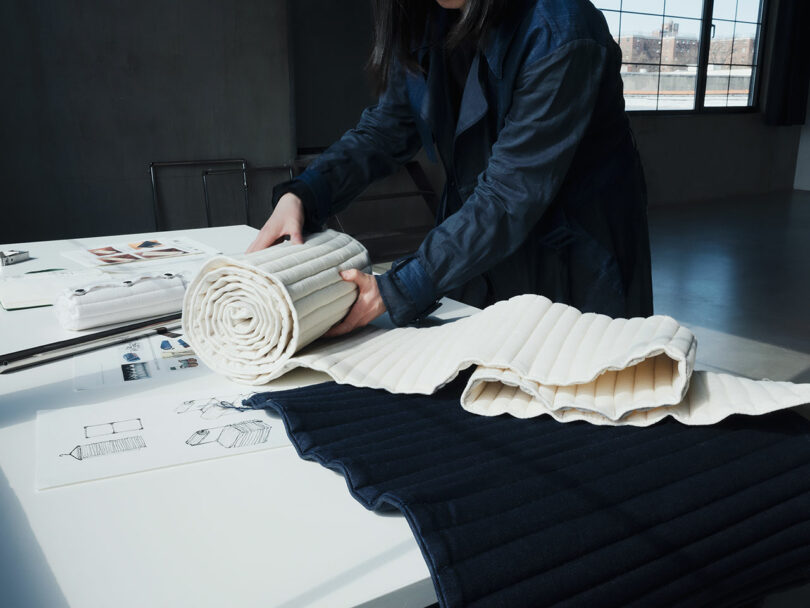 A person rolls up a ribbed textile sample on a table with fabric swatches and design sketches in a well-lit room.