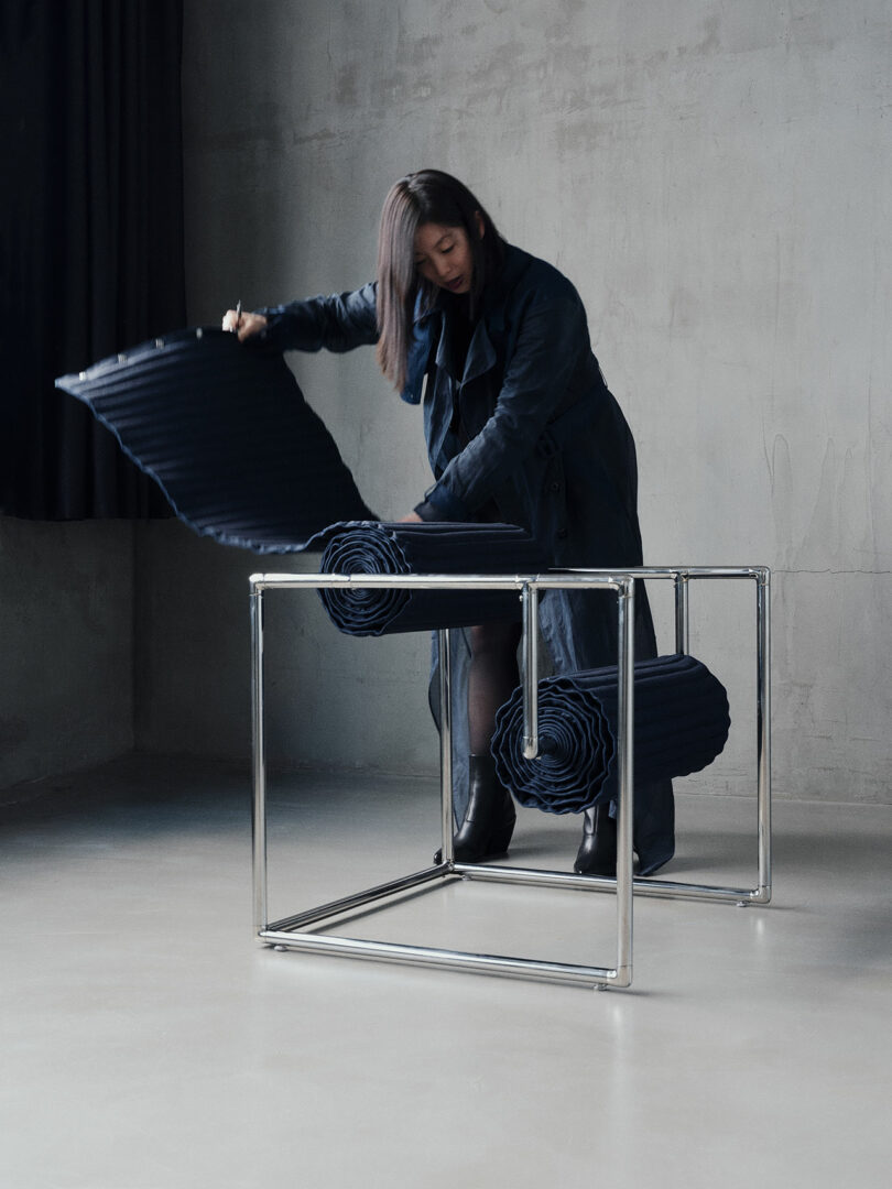 A woman adjusts a cushion on a modern chair with a metal frame and rolled fabric seating in a minimalist room with concrete walls and floors.