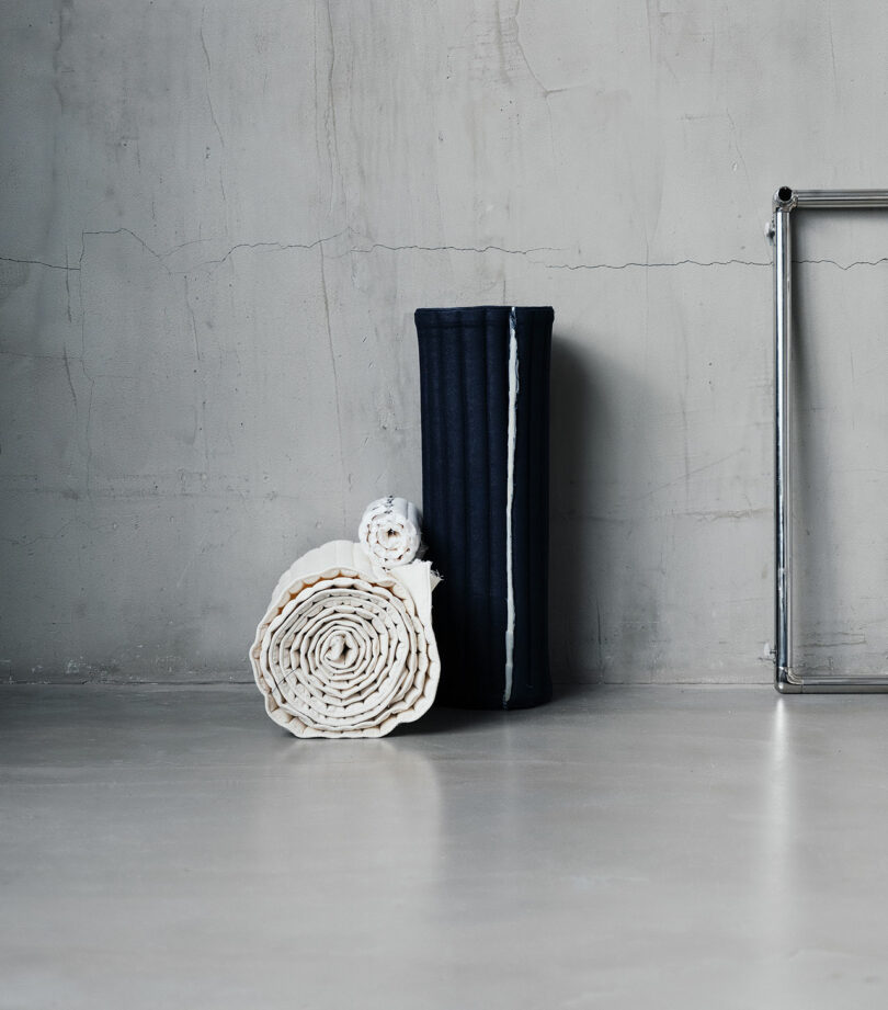 Two rolled and one folded textiles, one dark and two light-colored, displayed against a plain concrete wall on a smooth floor.