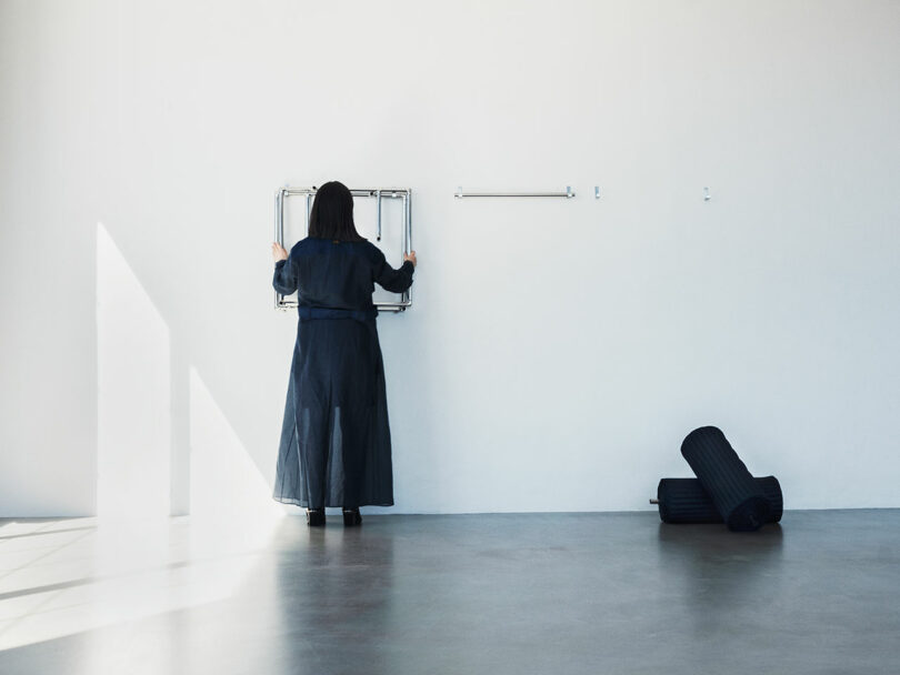 A person in a long dark coat faces a white wall, interacting with a foldable metal object; rolled-up dark mats rest on the floor nearby.