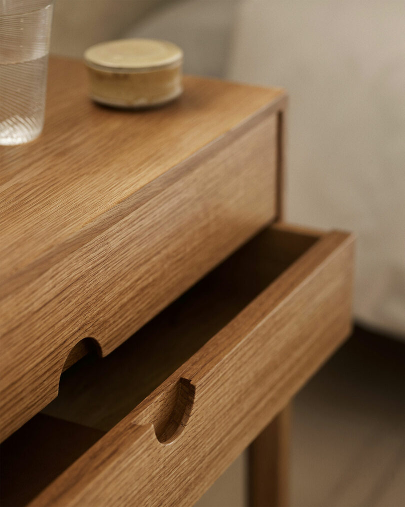 A close-up view of a wooden Entre nightstand with an open drawer, a glass of water, and a small round container on top.