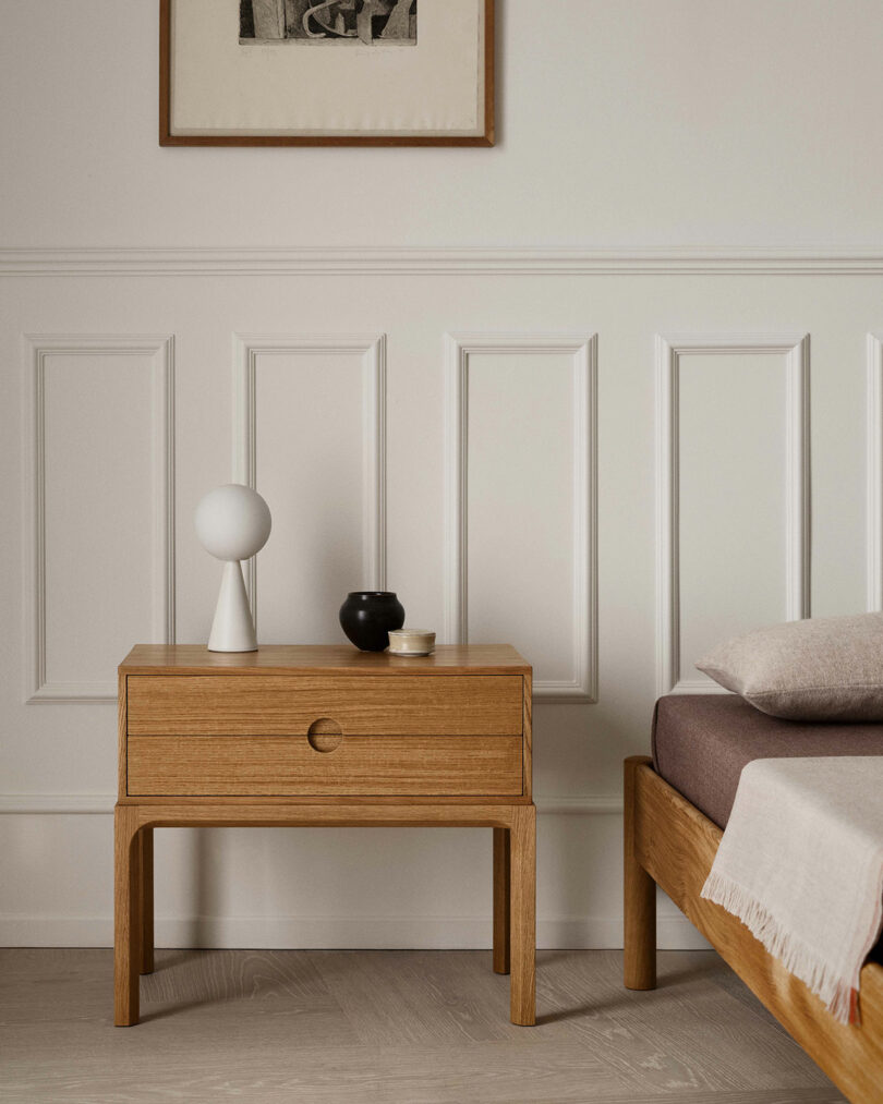 A wooden nightstand with a round lamp, a small black vase, and two stacked dishes—reminiscent of Entre Kai Kristiansen design—sits beside a bed with neutral bedding against a white-paneled wall.