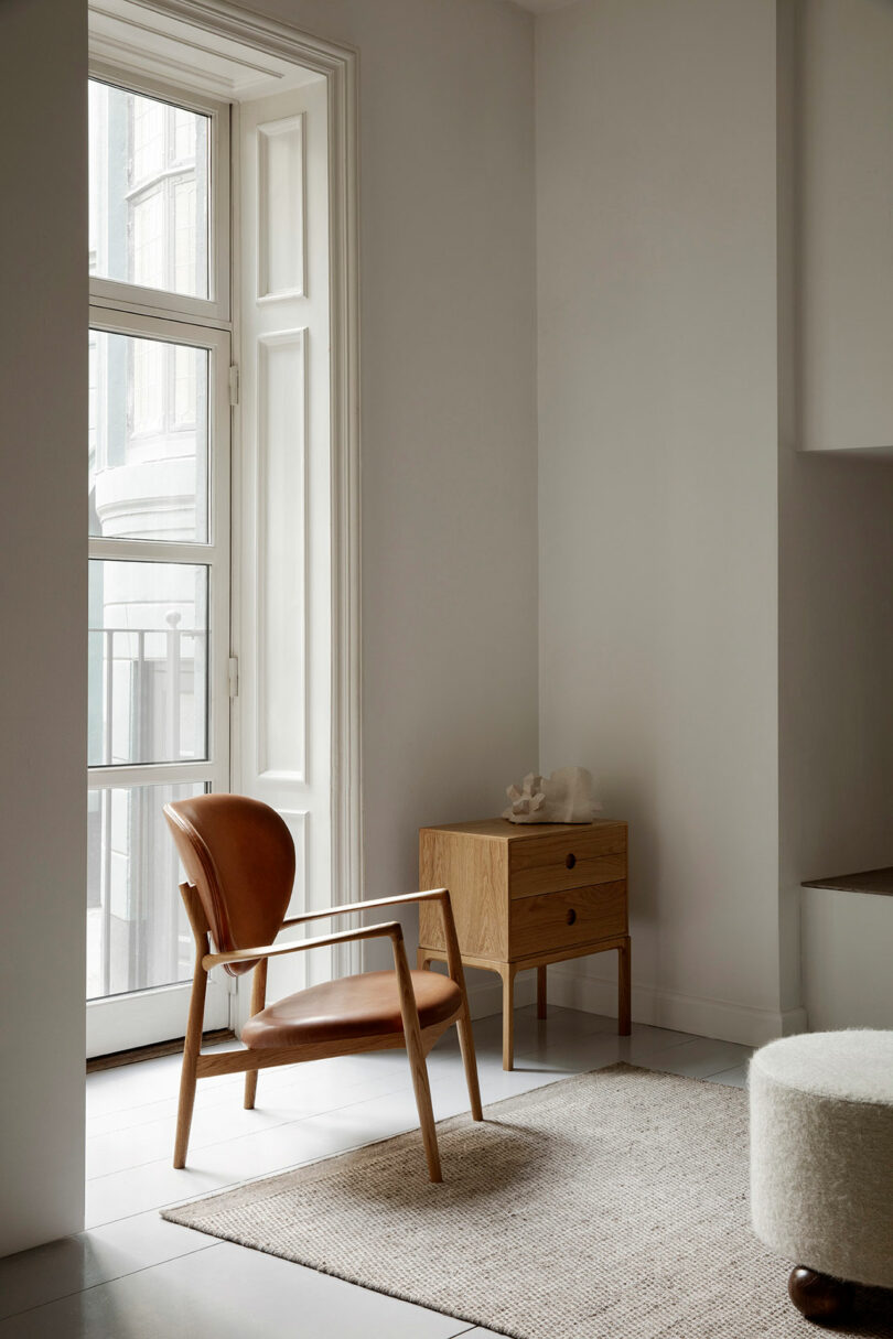 A minimalist room with a brown leather chair, a wooden Entre side table, a beige rug, and a large window letting in natural light.