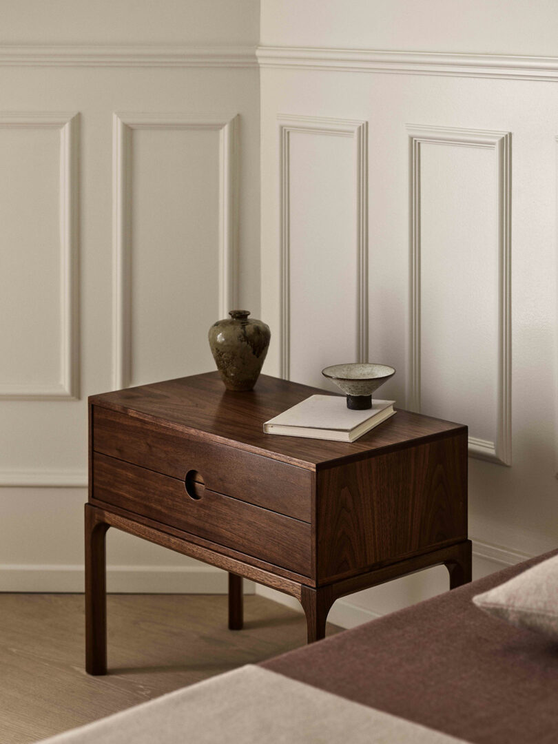 A wooden nightstand by Entre holds a ceramic vase, a bowl, and a closed book in Dagmar style, positioned next to a bed against a paneled wall.
